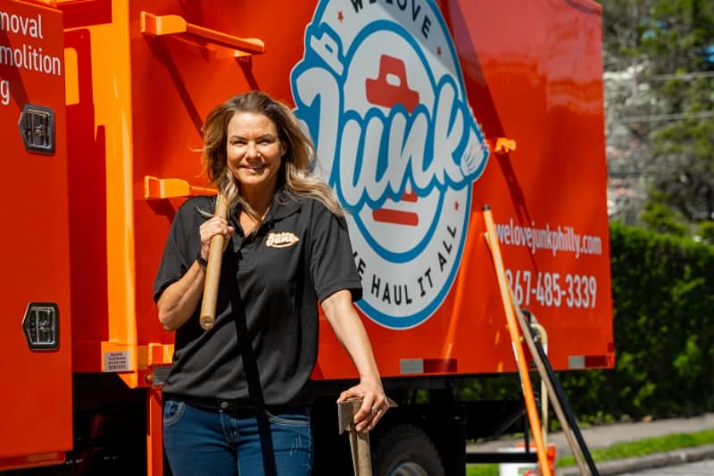 a women standing while holding a hammer and other tools beside her in front of a junk removal service truck